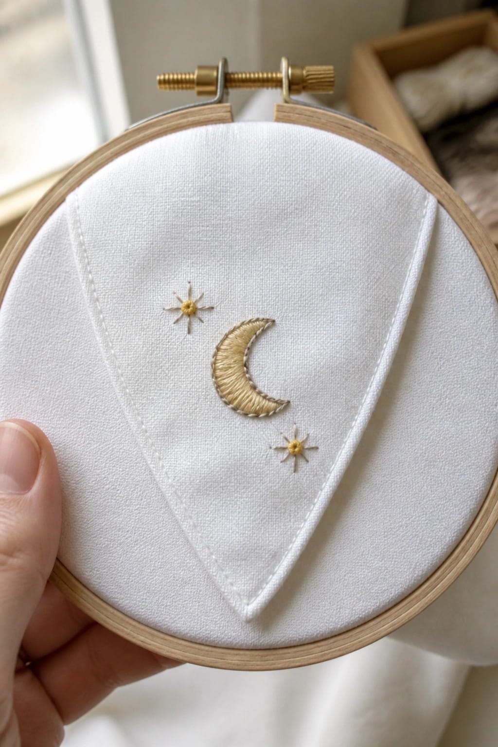 Hand holding an embroidery hoop with white fabric showing a stitched crescent moon and two stars on a triangular section.