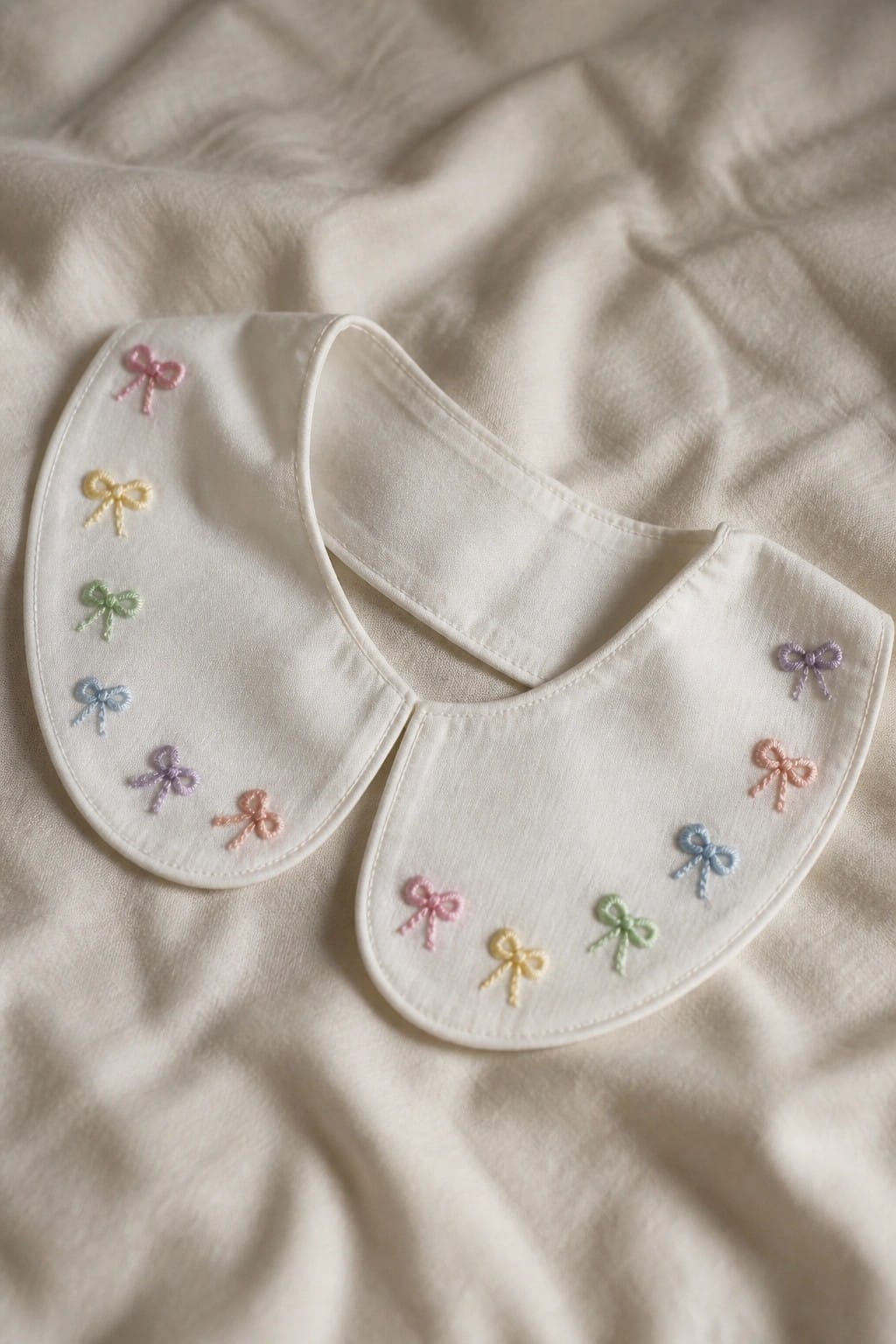 White bib with pastel rainbow bow embroidery on textured fabric surface