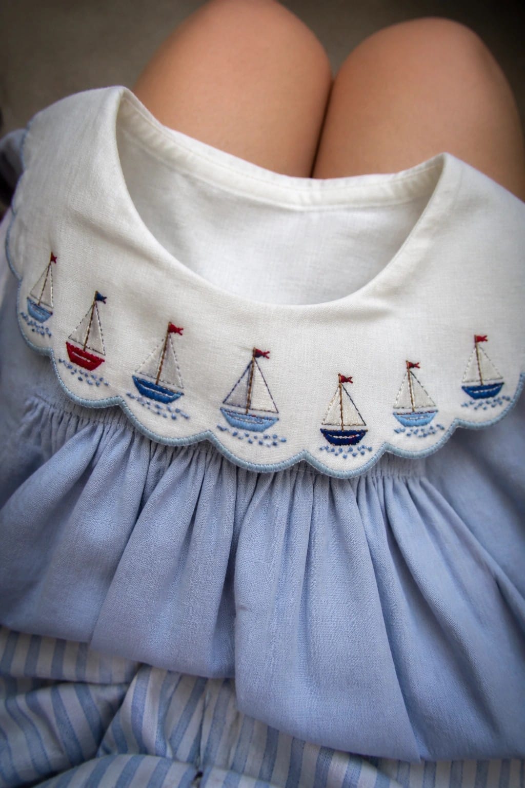 Close-up of a white scalloped collar on a light blue garment with embroidered sailboats in blue, red, and white thread.