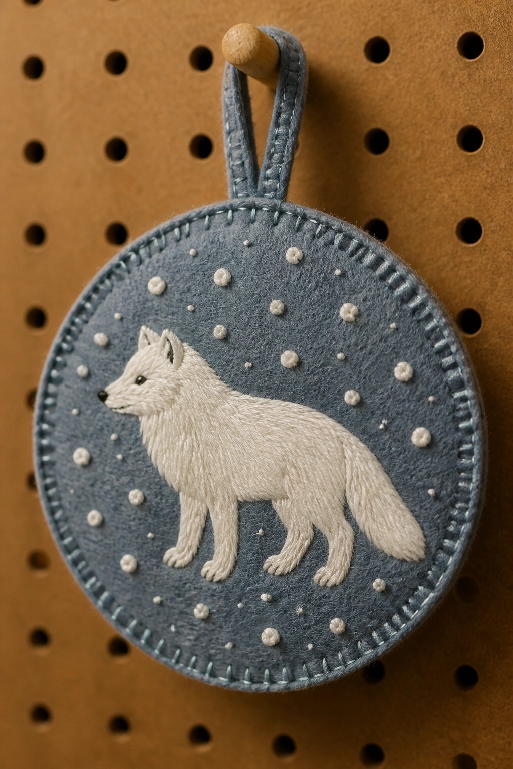 A circular blue felt piece with an embroidered white fox and scattered white dots hangs from a fabric loop on a pegboard.