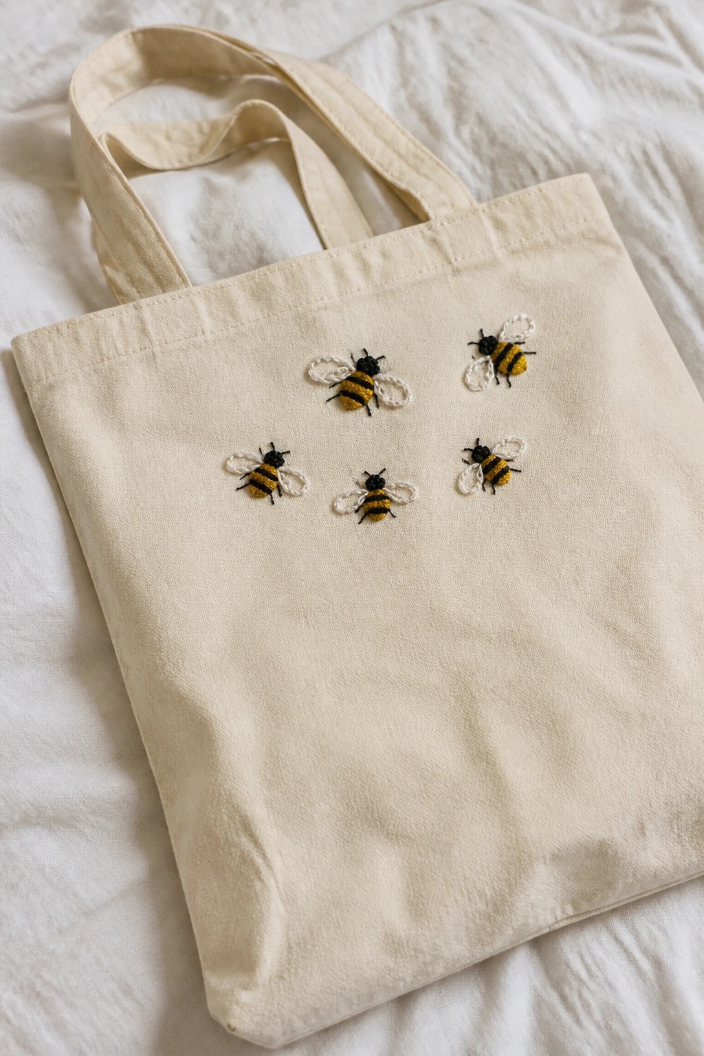 A cream canvas tote bag with five small embroidered bees in yellow and black scattered across the front.