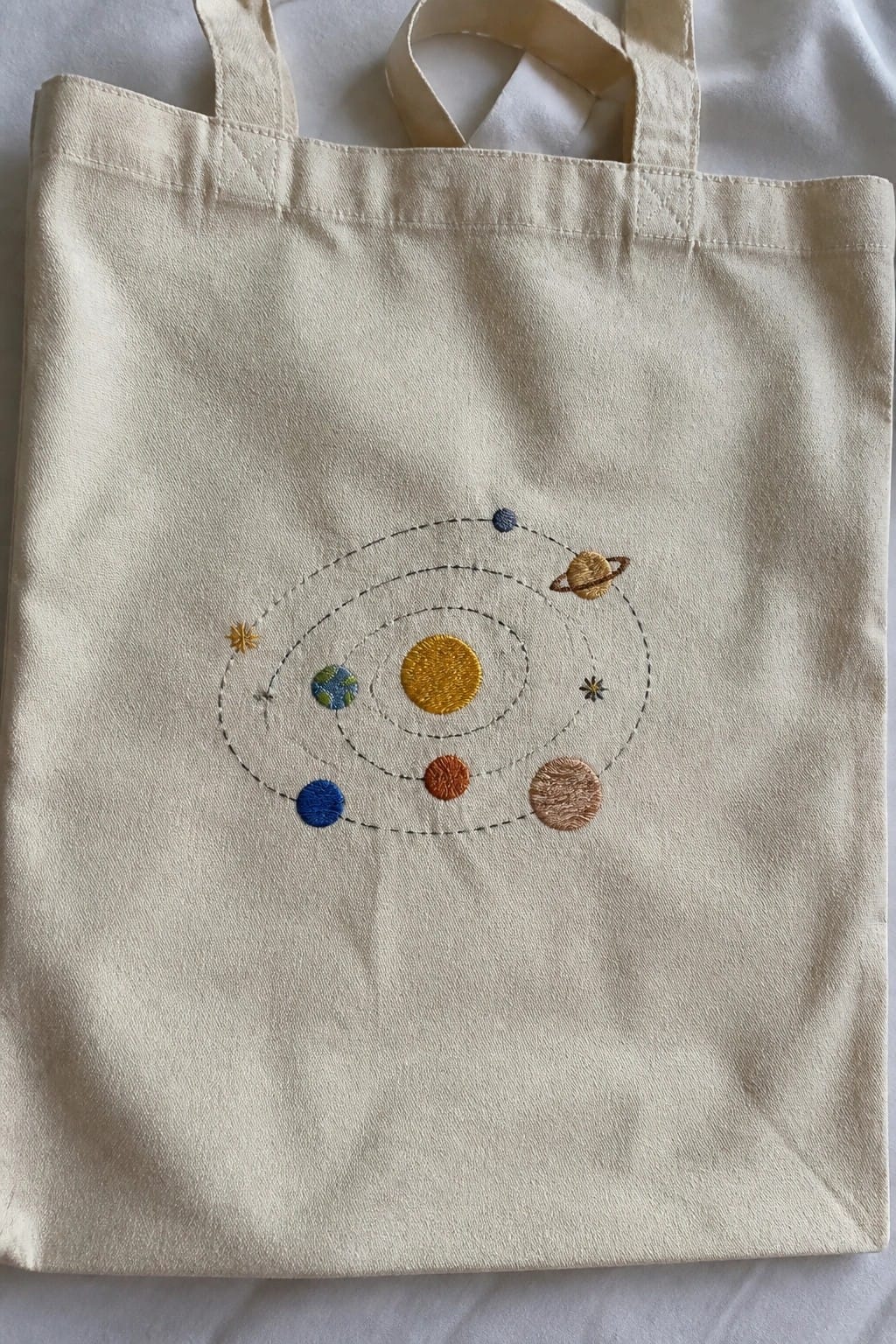 A canvas tote bag with an embroidered solar system showing a yellow sun, several planets, dashed orbital lines, and small stars.