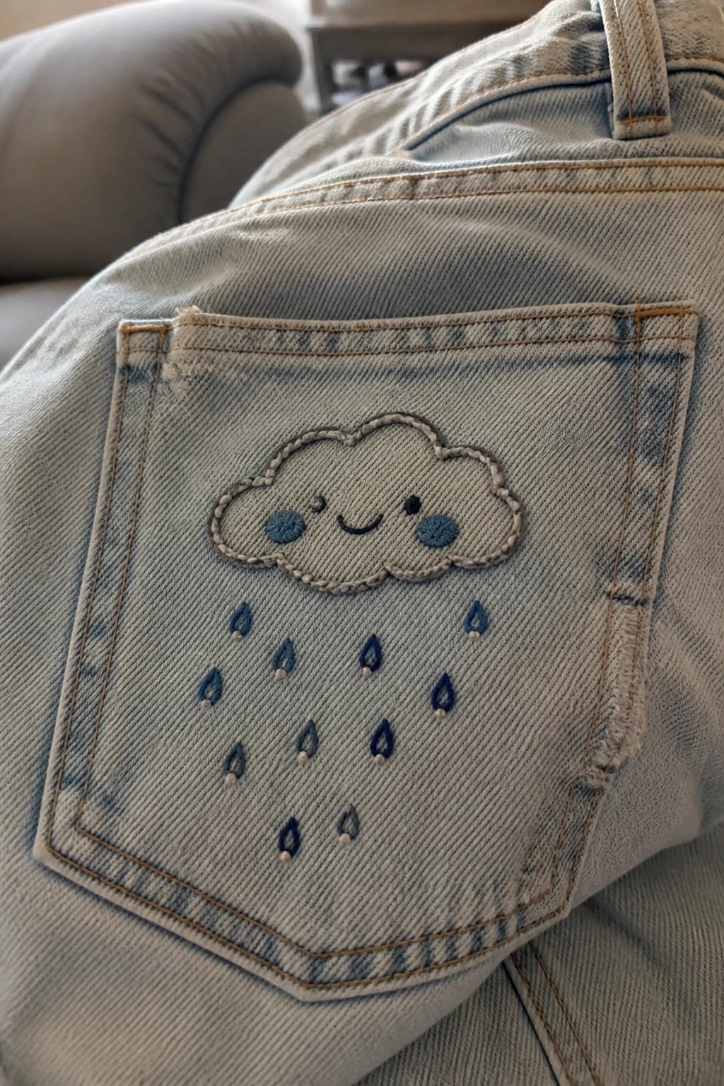 Light-wash denim jeans pocket with a blue embroidered cloud face and scattered raindrops.