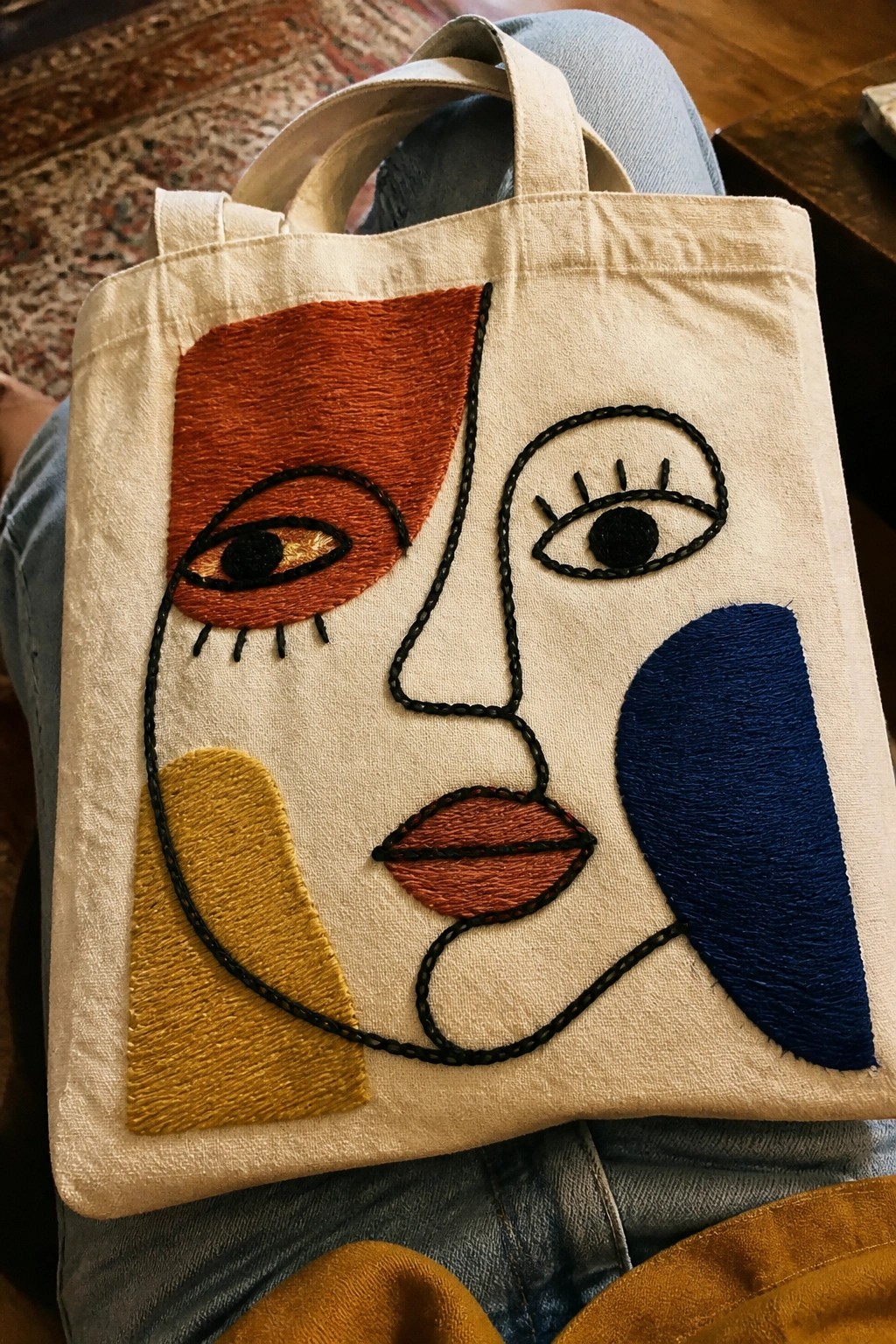 Beige tote bag with embroidered abstract face in orange, blue, and yellow