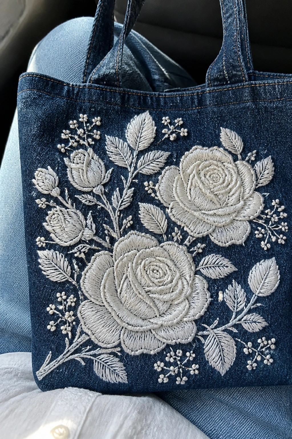 Blue denim tote bag with white embroidered roses, leaves, and small filler flowers on the front panel.