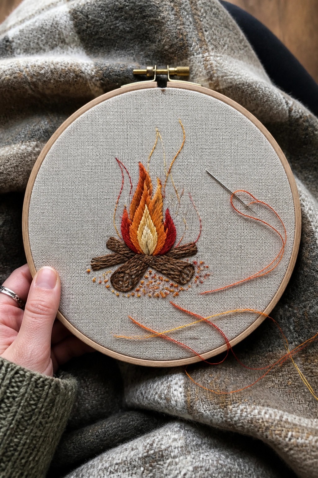 Hand holding a wooden embroidery hoop with a stitched campfire design on gray fabric and loose orange threads visible.