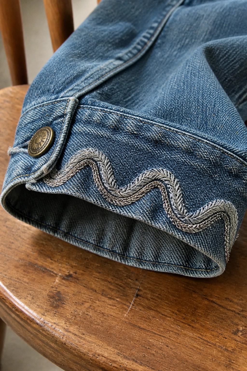 Blue denim cuff with wavy gray embroidery and brass button on wooden chair