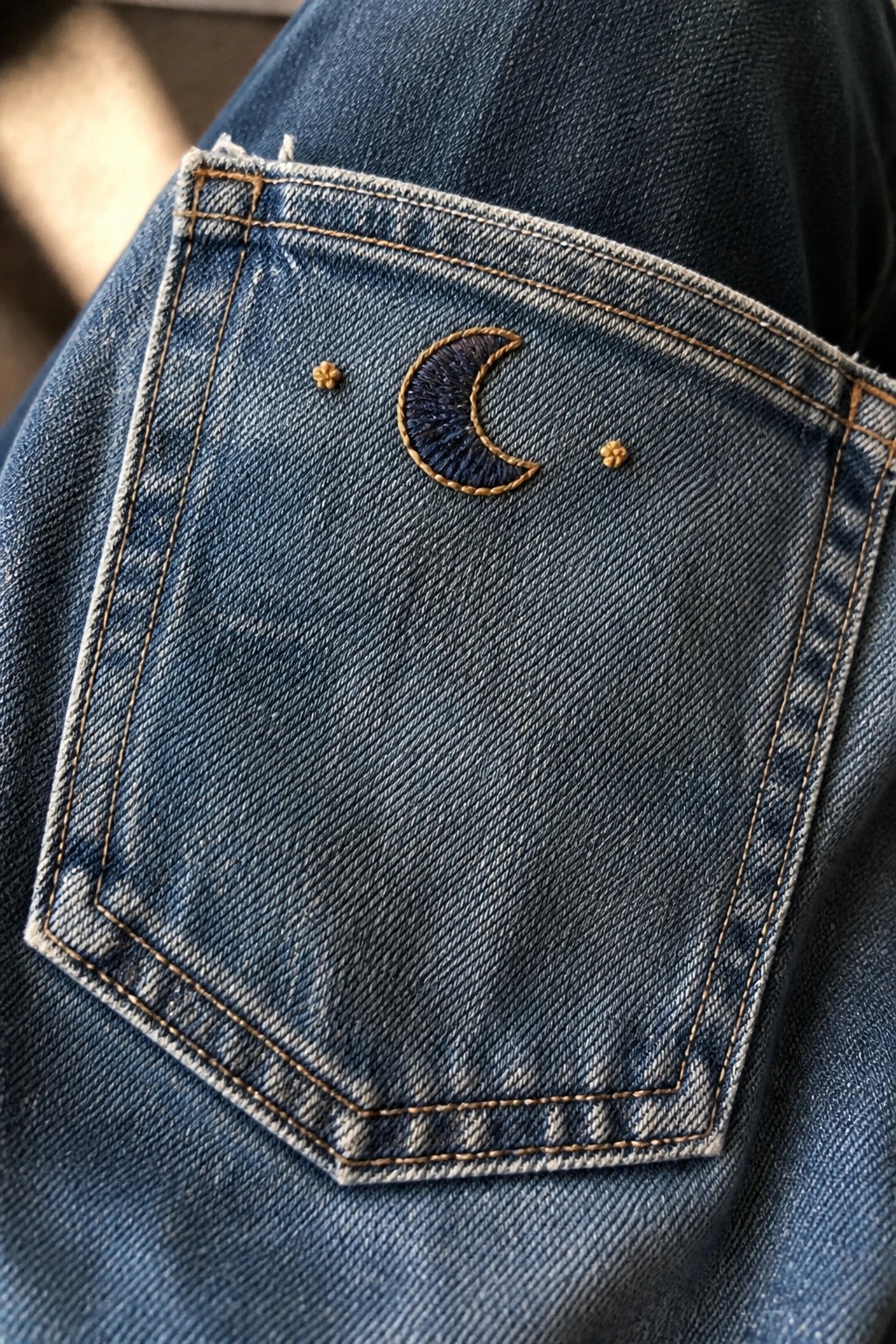 Close-up of blue denim jeans back pocket with dark blue crescent moon embroidery and two small gold stars on either side.