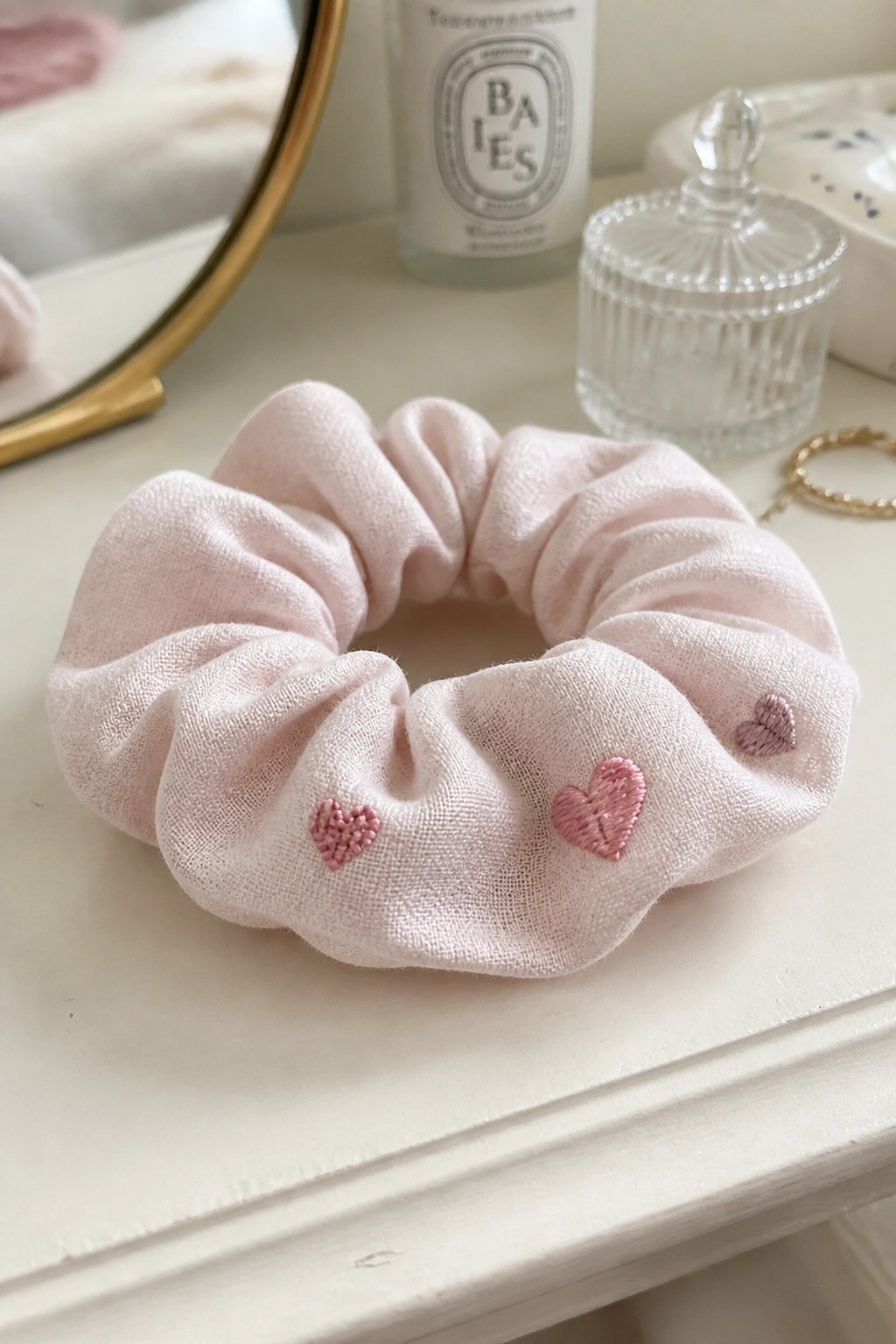 Light pink scrunchie with embroidered hearts on white vanity beside perfume bottle