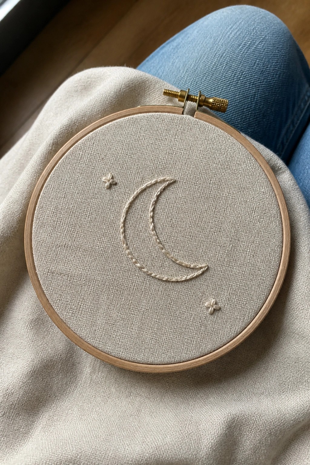 Wooden embroidery hoop with crescent moon and flowers on beige fabric over blue jeans.