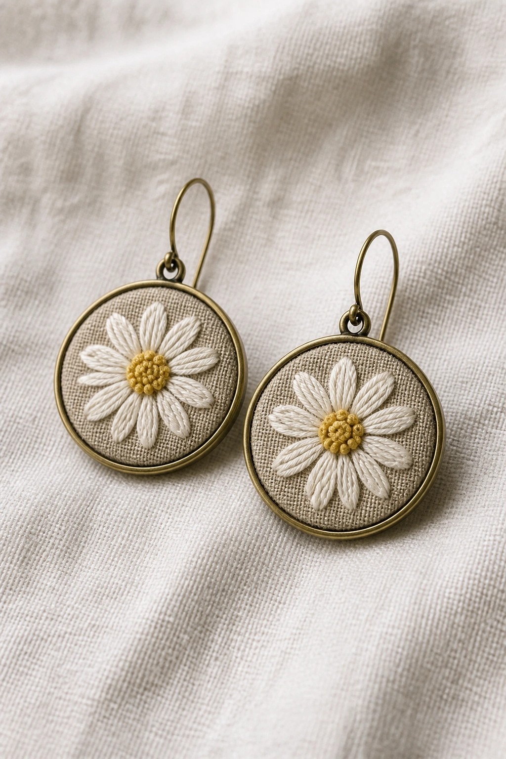 Two round brass hoop earrings with white daisy embroidery and yellow centers on beige fabric.