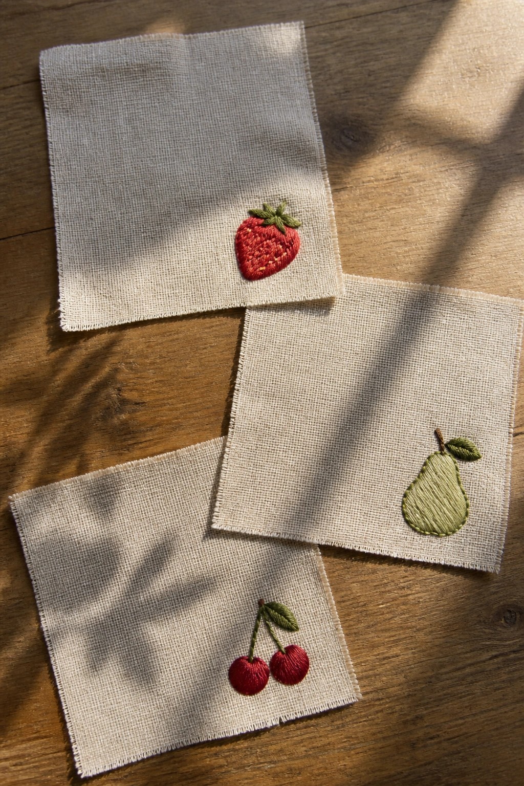 Three small off-white fabric squares on a wooden surface, each embroidered with a different fruit: a red strawberry, two red cherries, and a green pear.