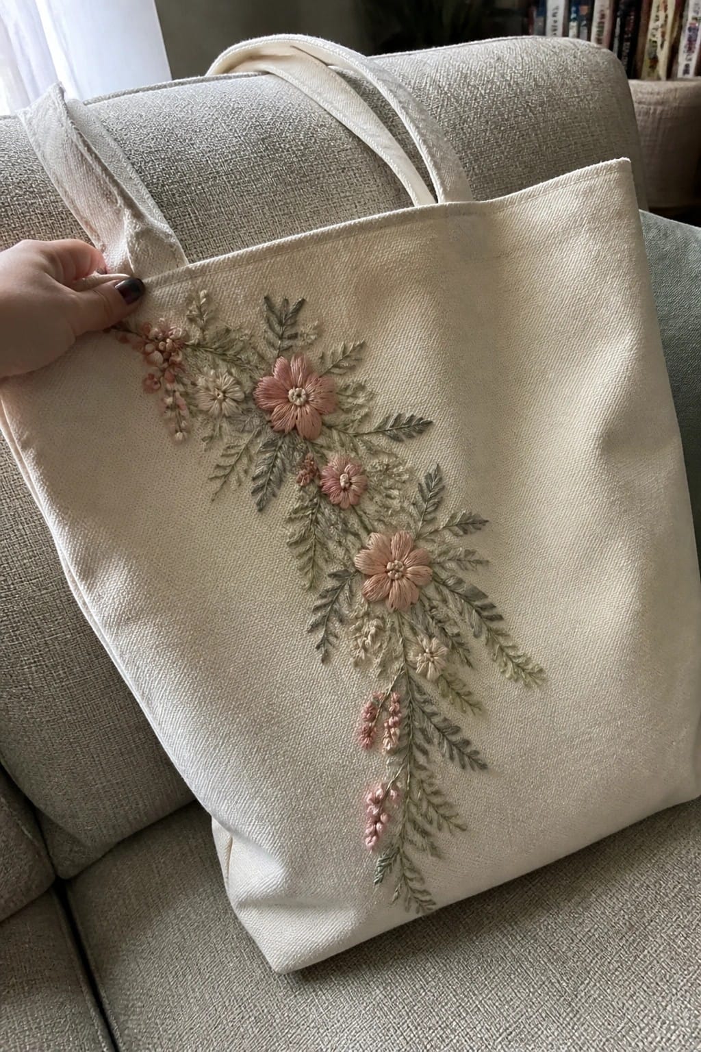A hand holds a cream canvas tote bag with pink and green floral embroidery resting on a couch.