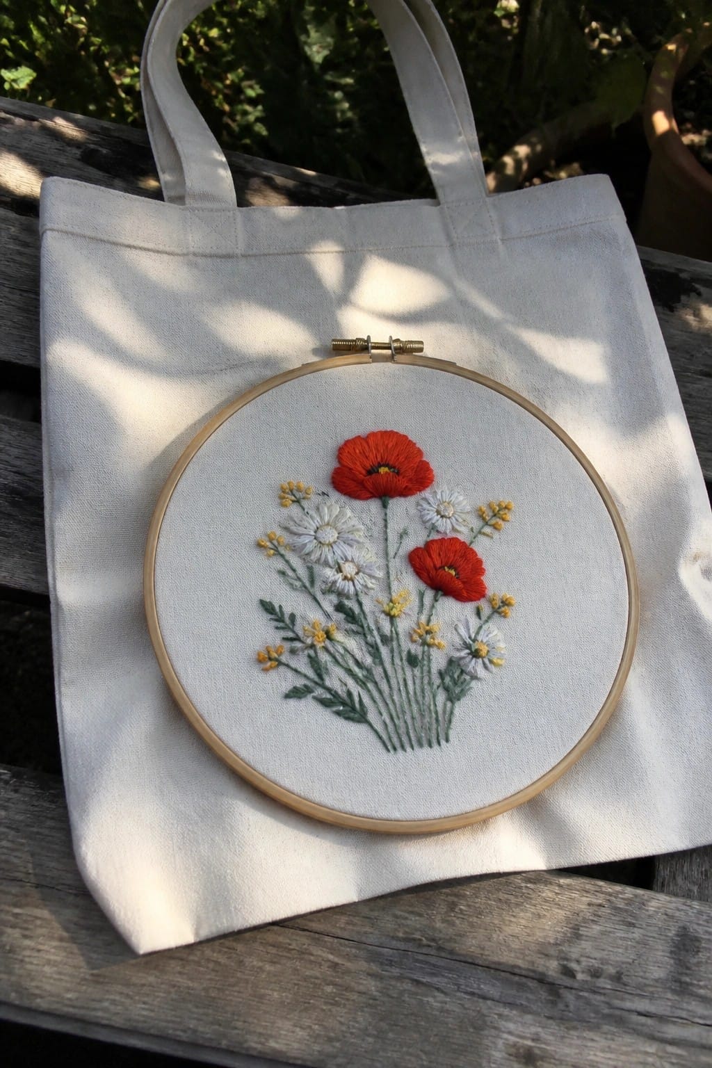 A white canvas tote bag with an embroidery hoop showing red poppies, white daisies, and yellow flowers stitched in the center.