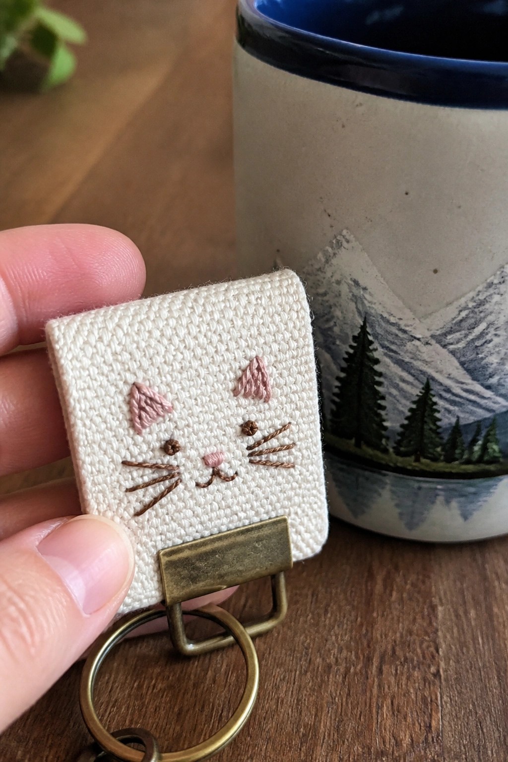 Hand holding embroidered white cat keychain near mountain-patterned ceramic mug.