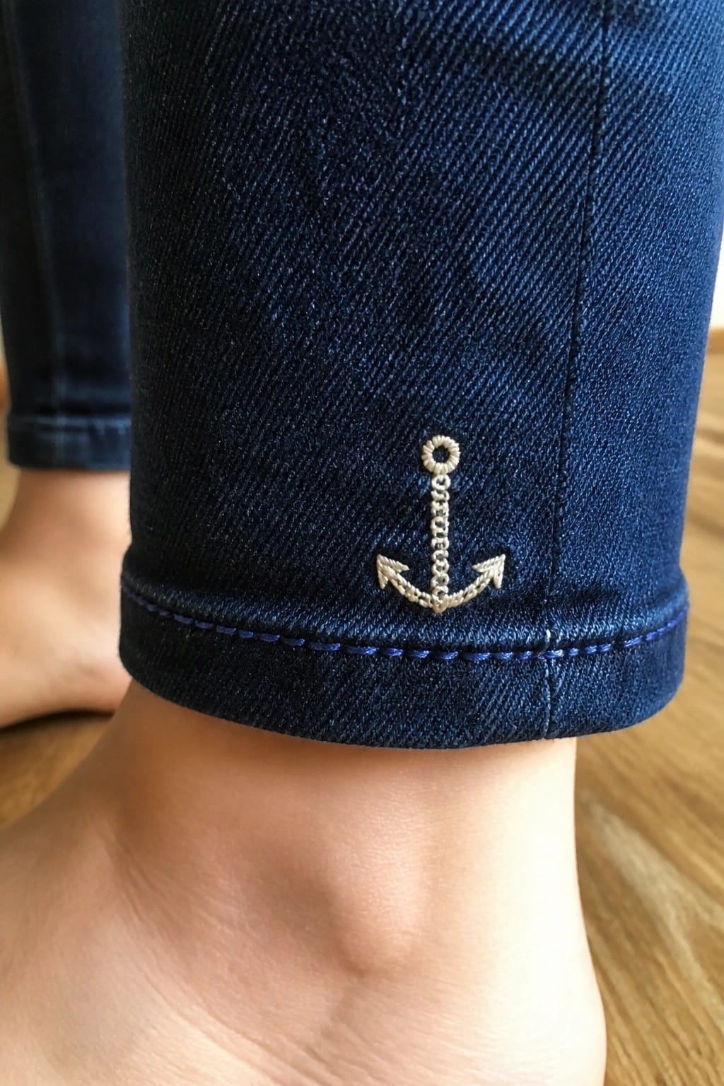 Dark blue cuffed denim jeans with embroidered anchor above bare ankle on wood floor