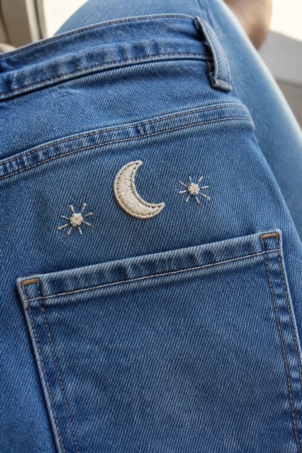 A close-up of a small red and black ladybug embroidered on the back pocket of blue denim jeans.