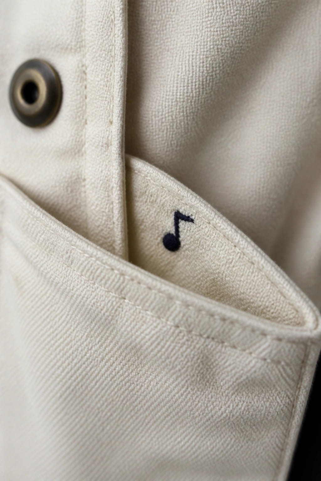 Cream jacket pocket with black musical note embroidery beside metal grommet