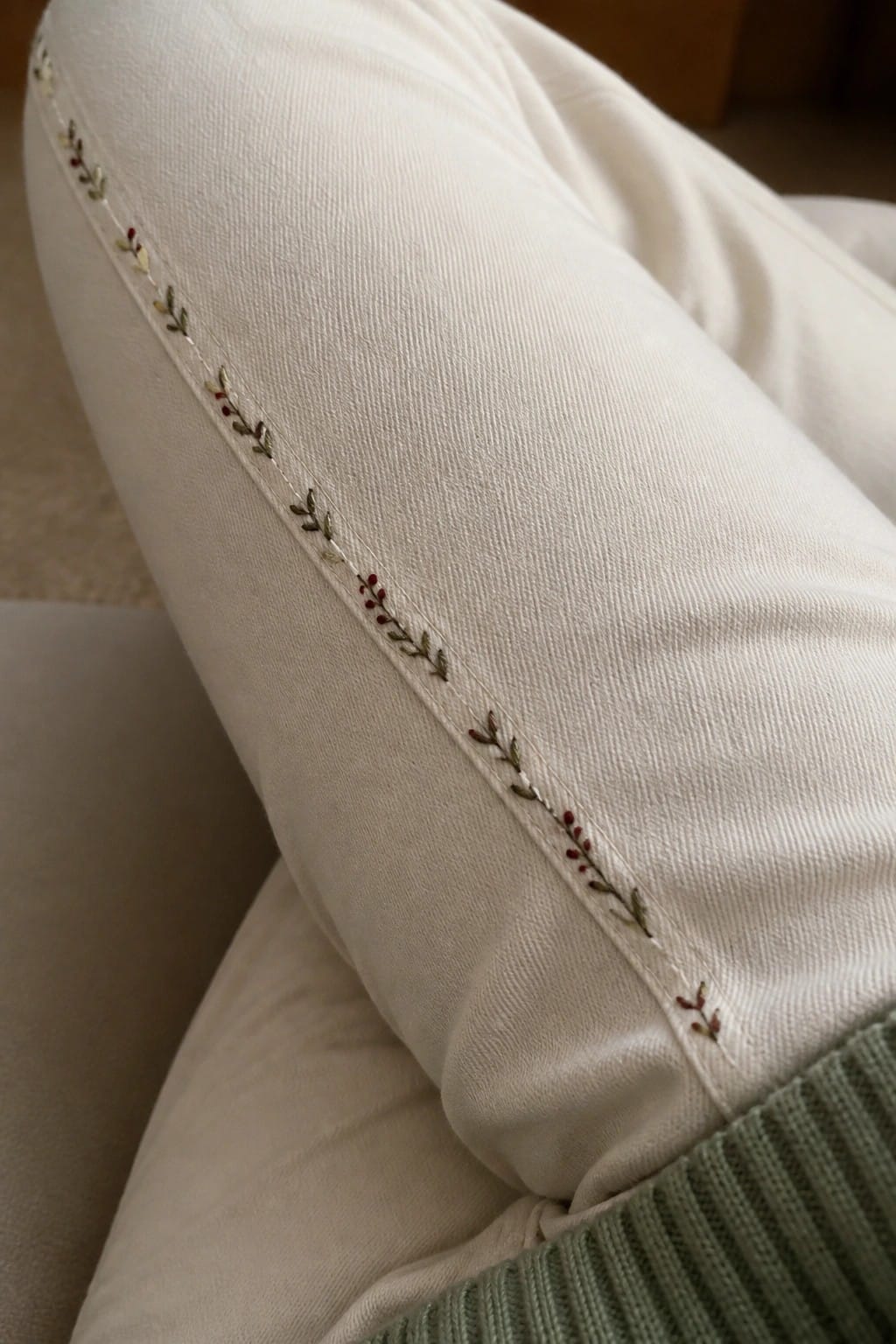 Close-up of white jeans showing a vertical line of small embroidered branches with green leaves and red dots along the side seam.