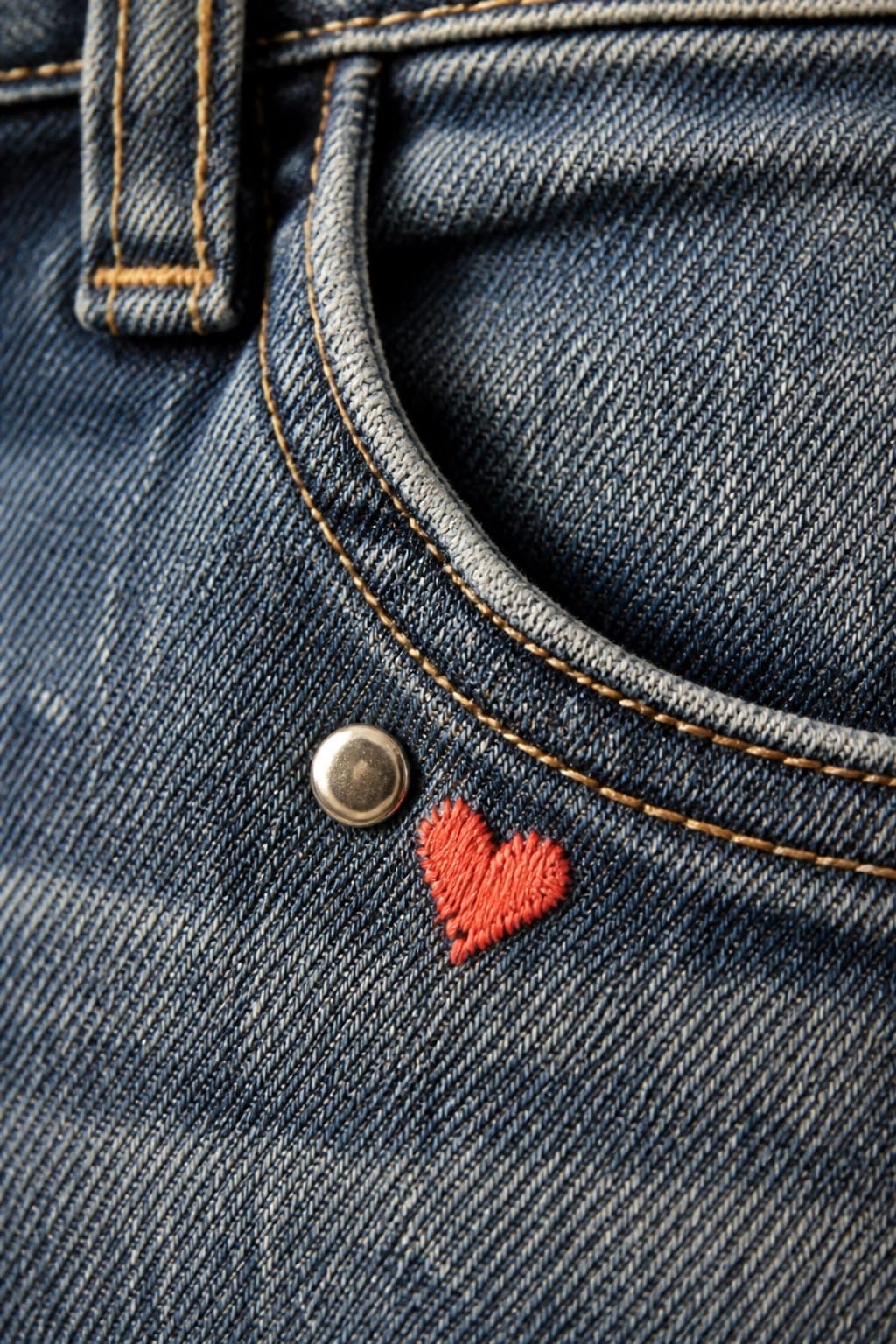 Blue denim jeans with red embroidered heart beside metal rivet and yellow stitching