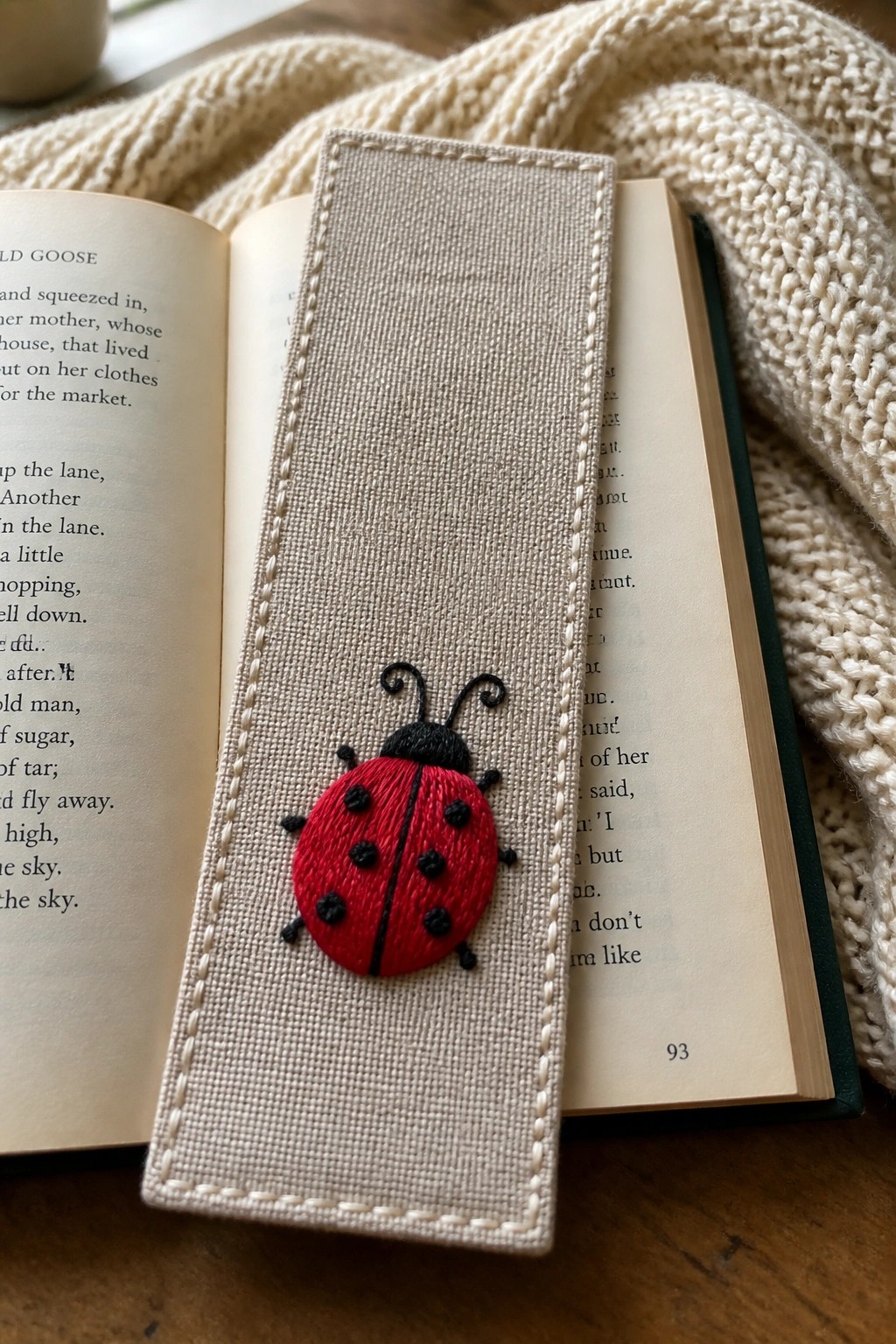 Open book with red embroidered ladybug on beige linen bookmark beside cream knit fabric.