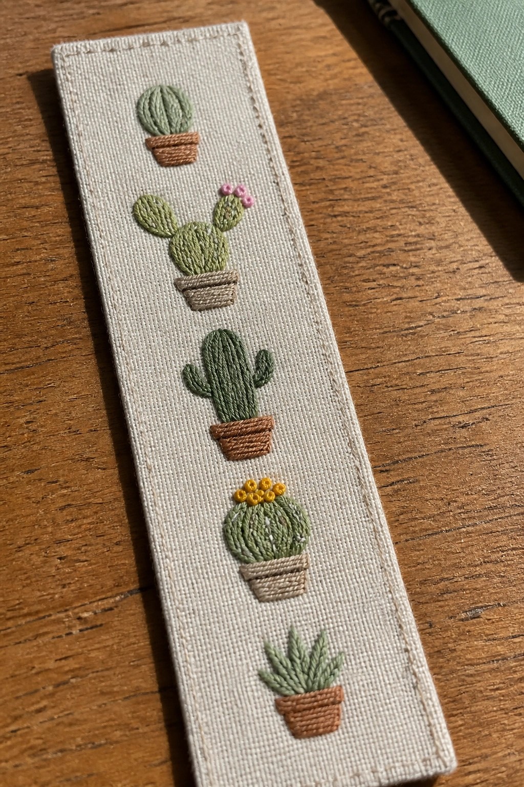 A narrow beige fabric bookmark with five embroidered potted cacti stacked vertically in different shapes and colors.