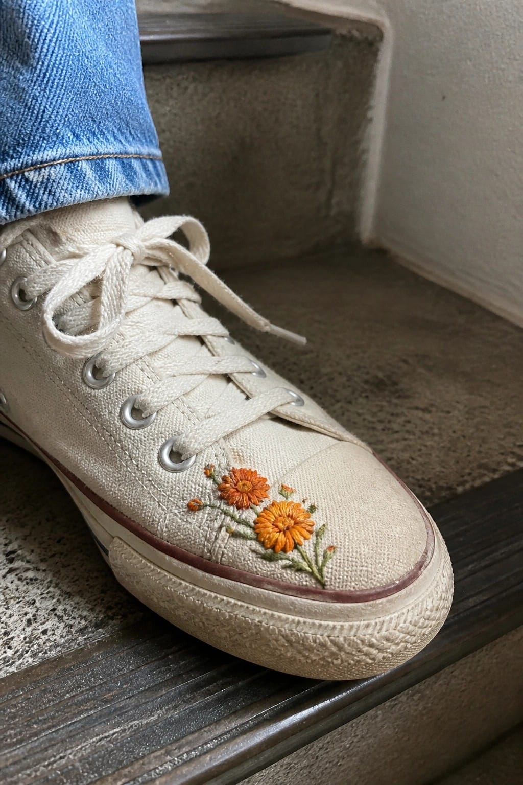 A white canvas sneaker with small orange embroidered flowers on the toe.