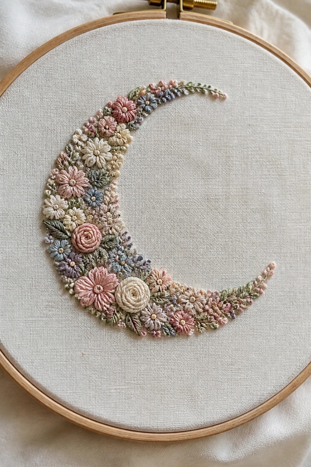 A wooden embroidery hoop with white fabric shows a crescent moon shape made of embroidered flowers in soft pastel colors.