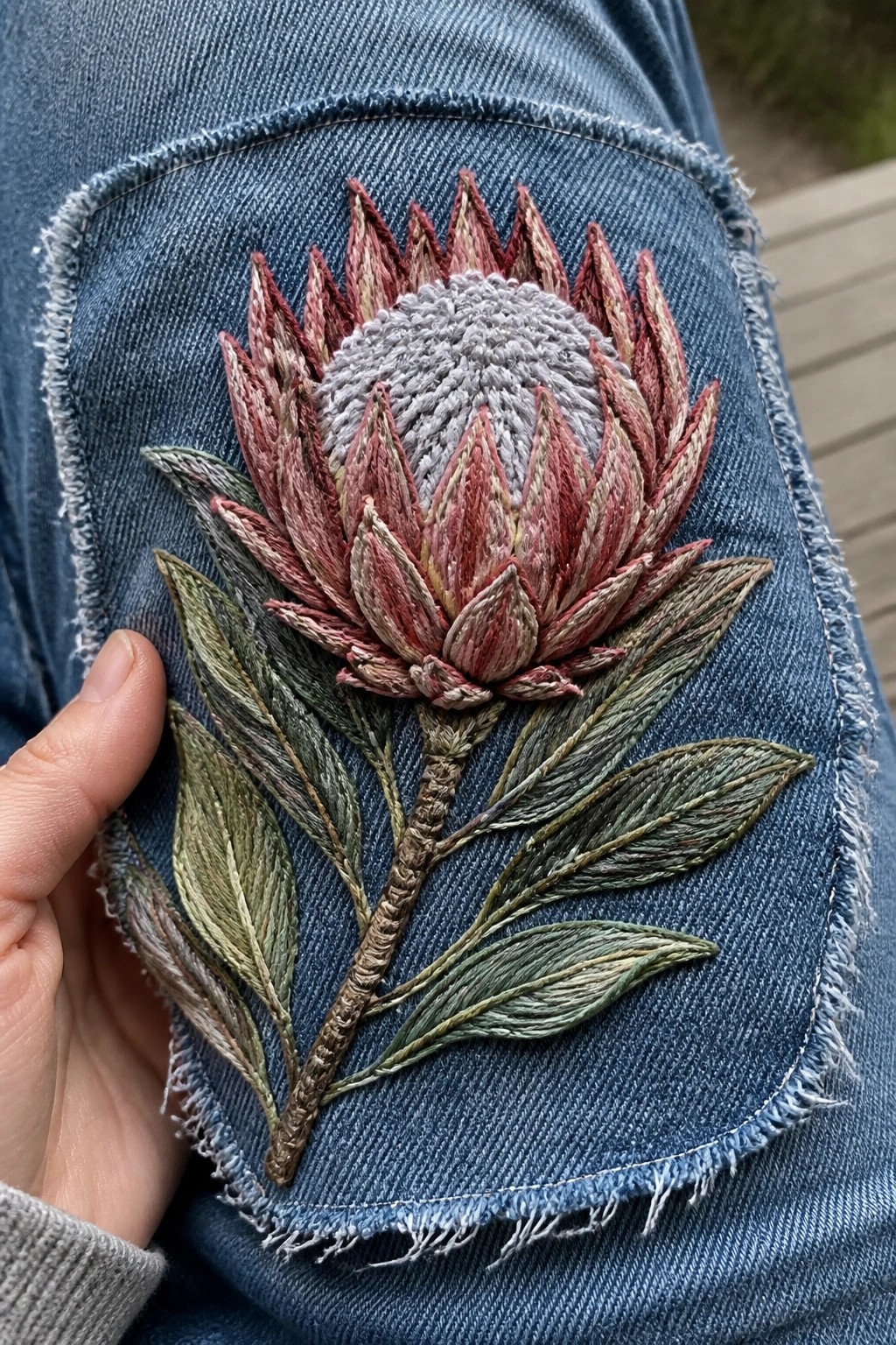 Close-up of a hand-embroidered protea flower with pink and gray threads on blue denim jeans fabric.