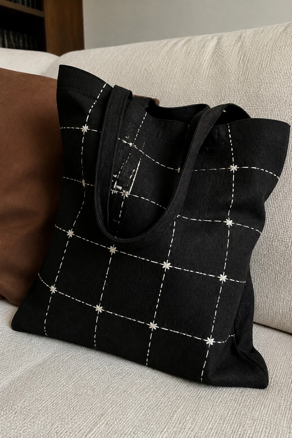 A black tote bag with white dashed grid lines and small star stitches at the intersections sits on a light couch.
