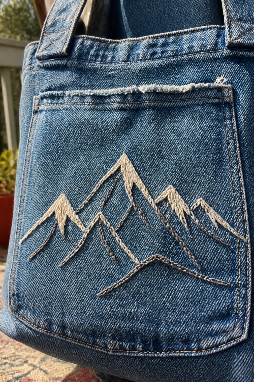 Close-up of blue denim tote bag pocket with white and gray embroidered mountain silhouettes.