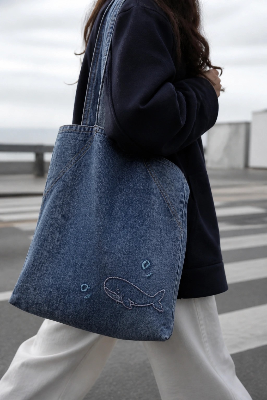 A blue denim tote bag with a stitched whale outline and small bubble circles carried on someone's shoulder.