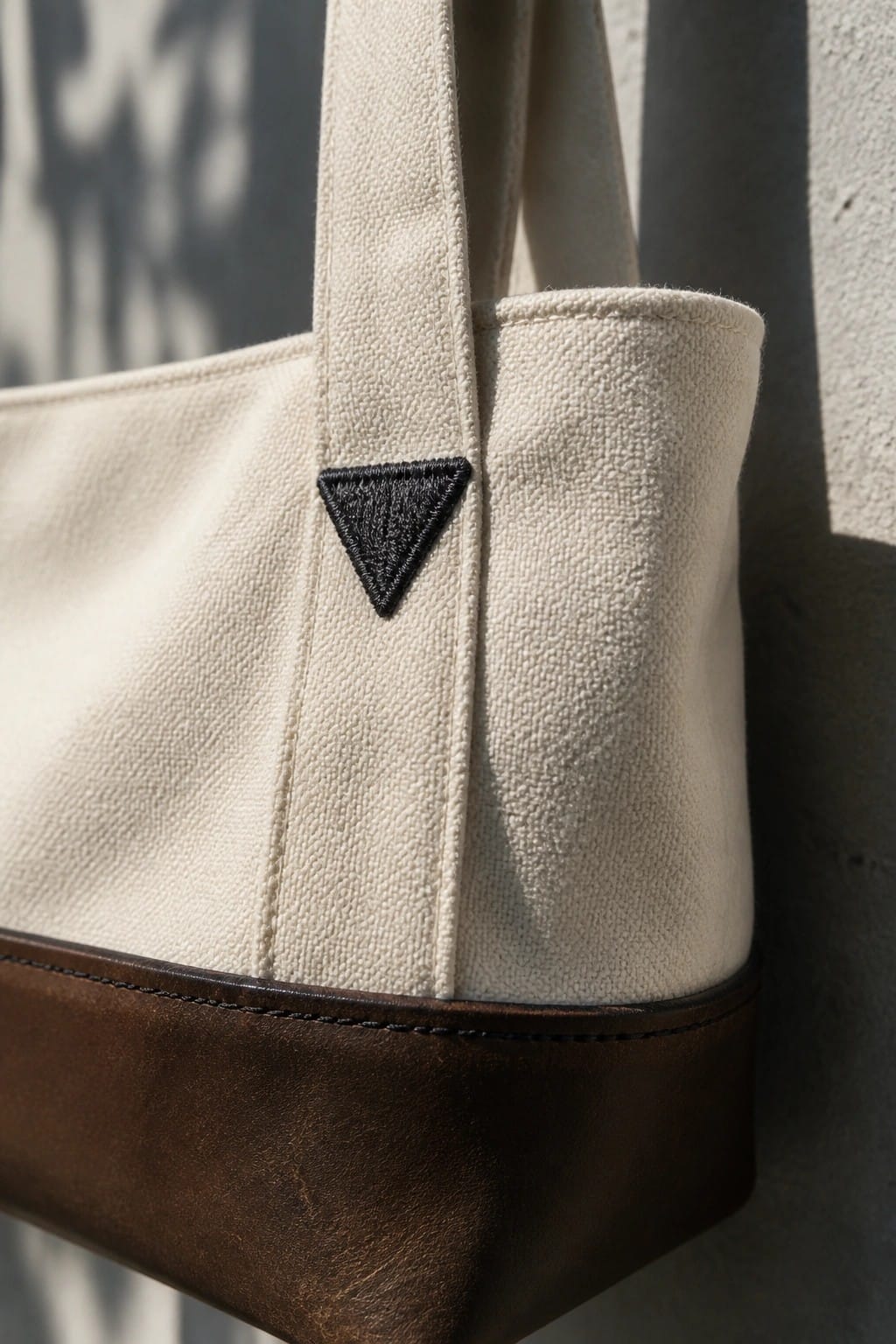 Close-up of a beige canvas tote bag showing a small black embroidered triangle near the top edge above a brown leather base.