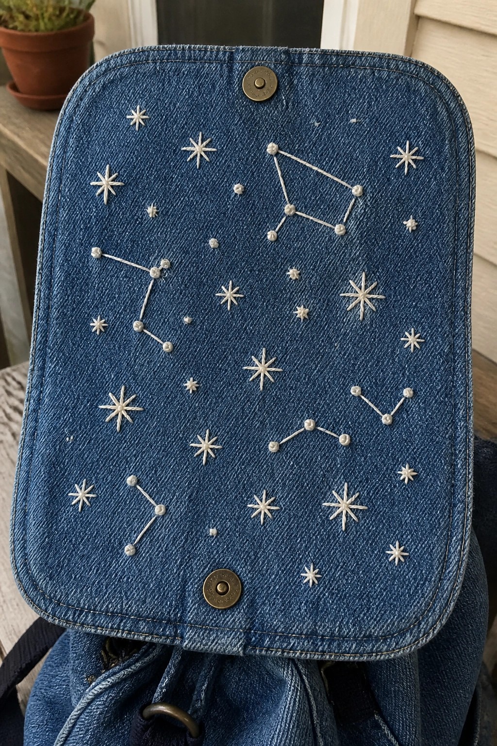 Blue denim bag flap embroidered with white stars and constellations, metal snap buttons.