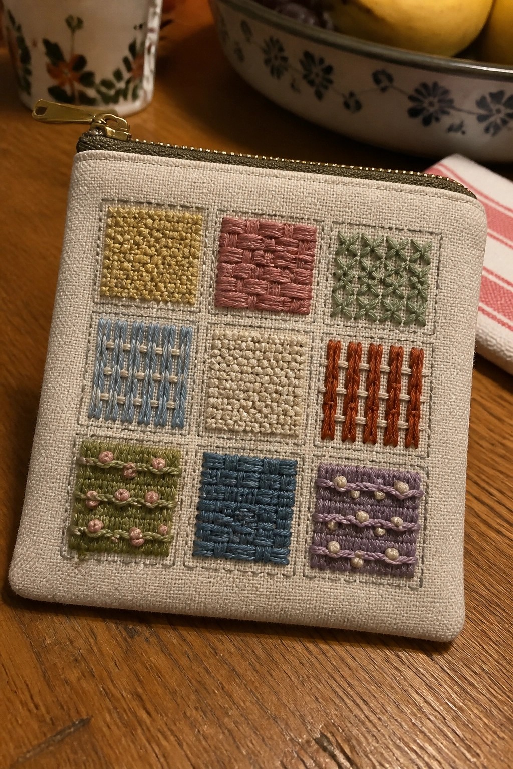 A small beige zippered pouch displays a 3x3 grid of embroidered squares in various colors and stitch patterns.