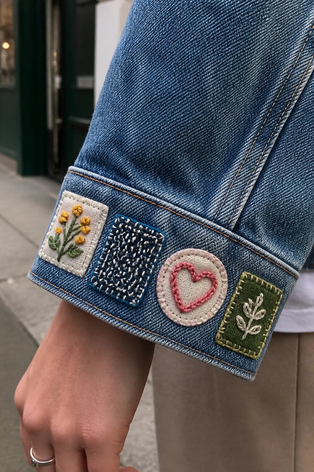Denim jacket cuff with embroidered patches of yellow flowers, pink heart, and green leaf