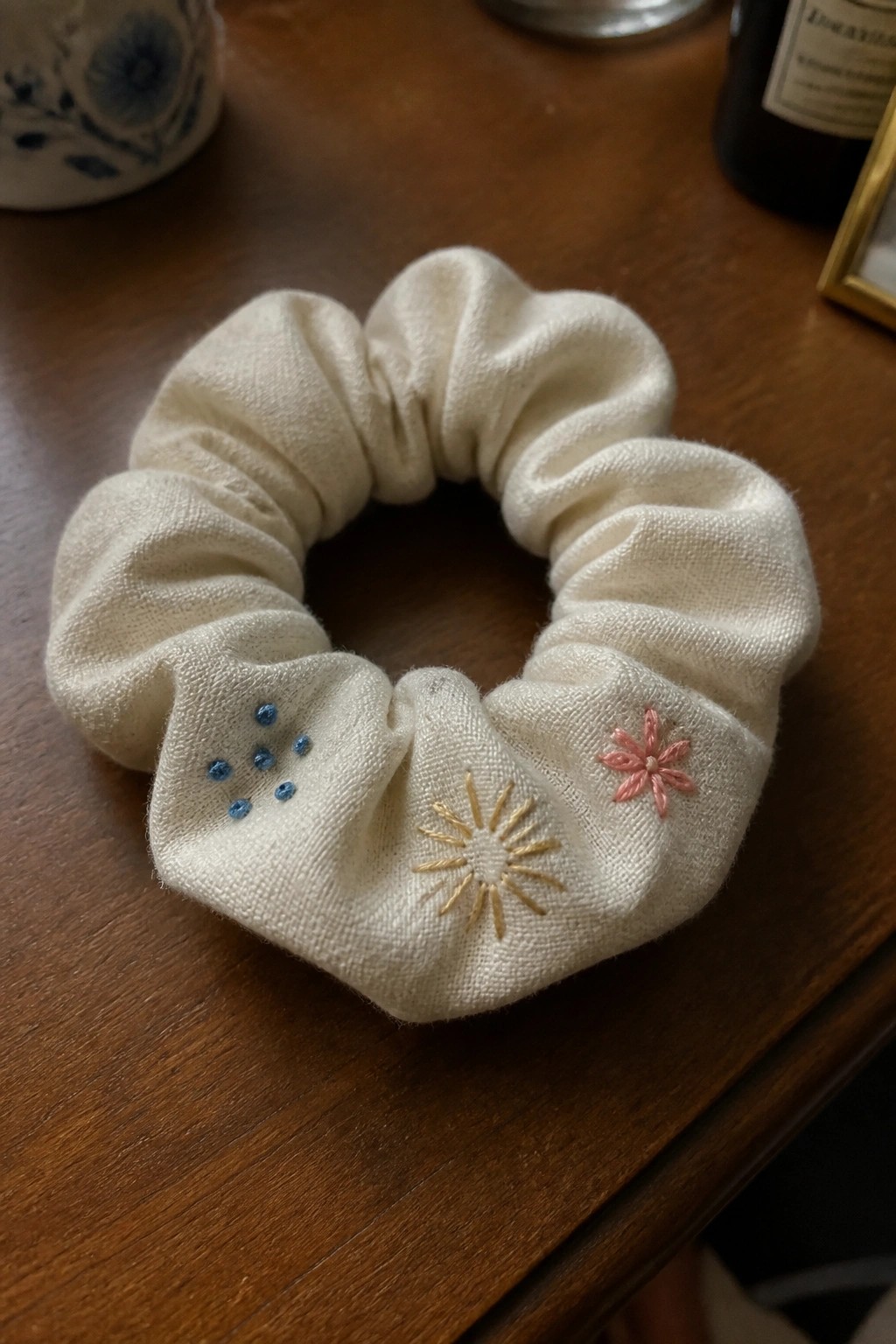 A white fabric scrunchie with blue dots, a yellow sunburst, and a pink flower embroidered on it resting on a wooden table.