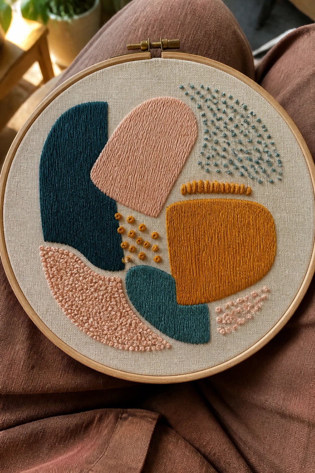 Abstract embroidery hoop with teal, peach, and mustard shapes on beige fabric over brown lap.