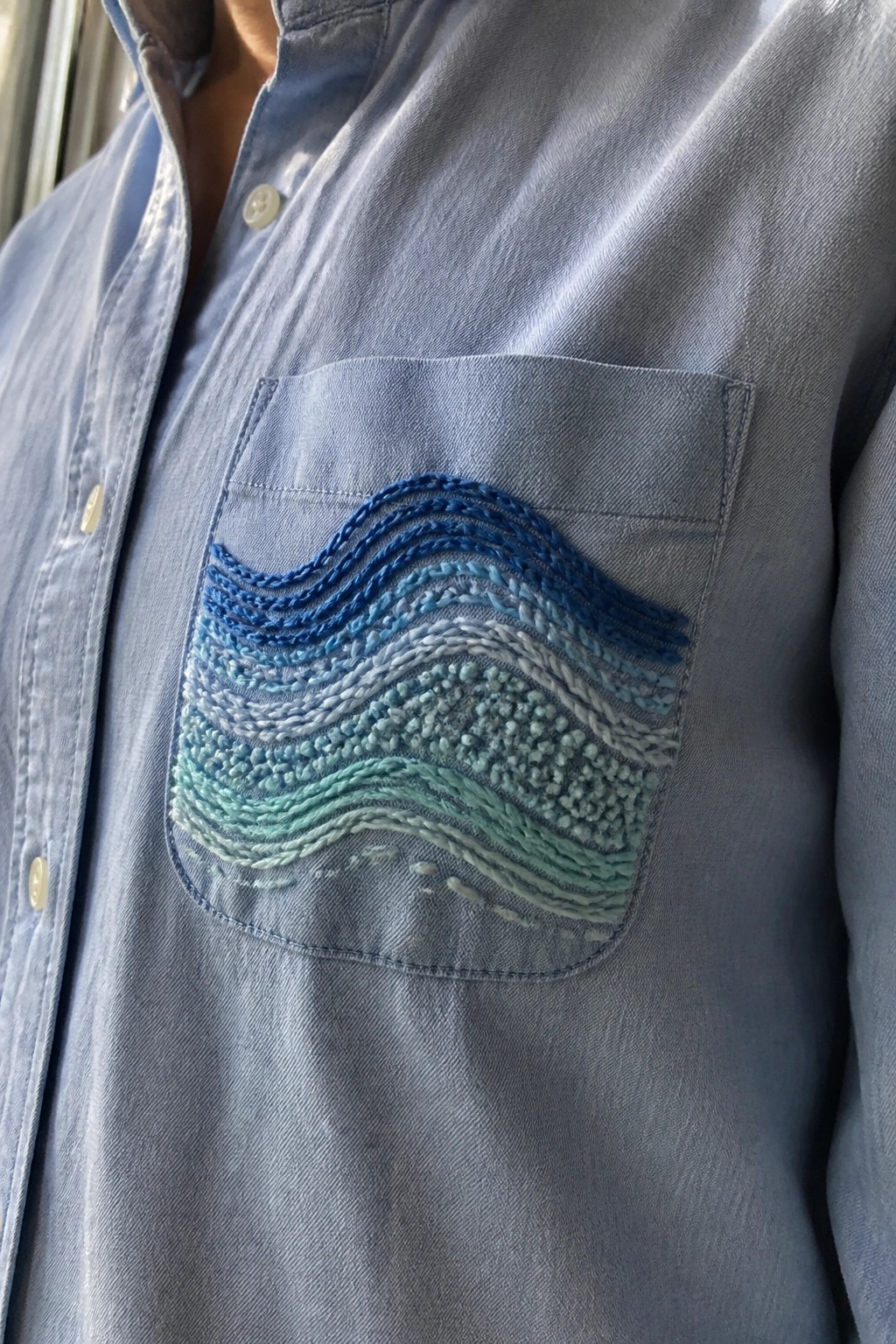 Blue button-up shirt with a chest pocket covered in horizontal wavy embroidery lines in shades of blue and green.