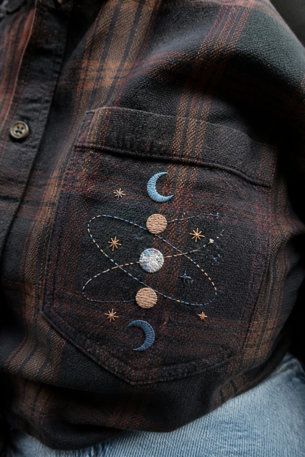 Close-up of celestial embroidery with moons, planets, stars, and orbit lines on the pocket of a brown plaid shirt.