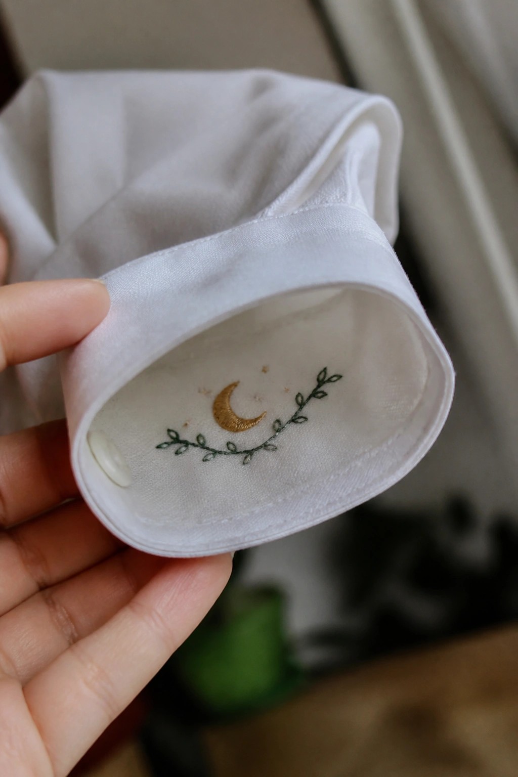 Hand holding white shirt cuff with gold moon and green vine embroidery