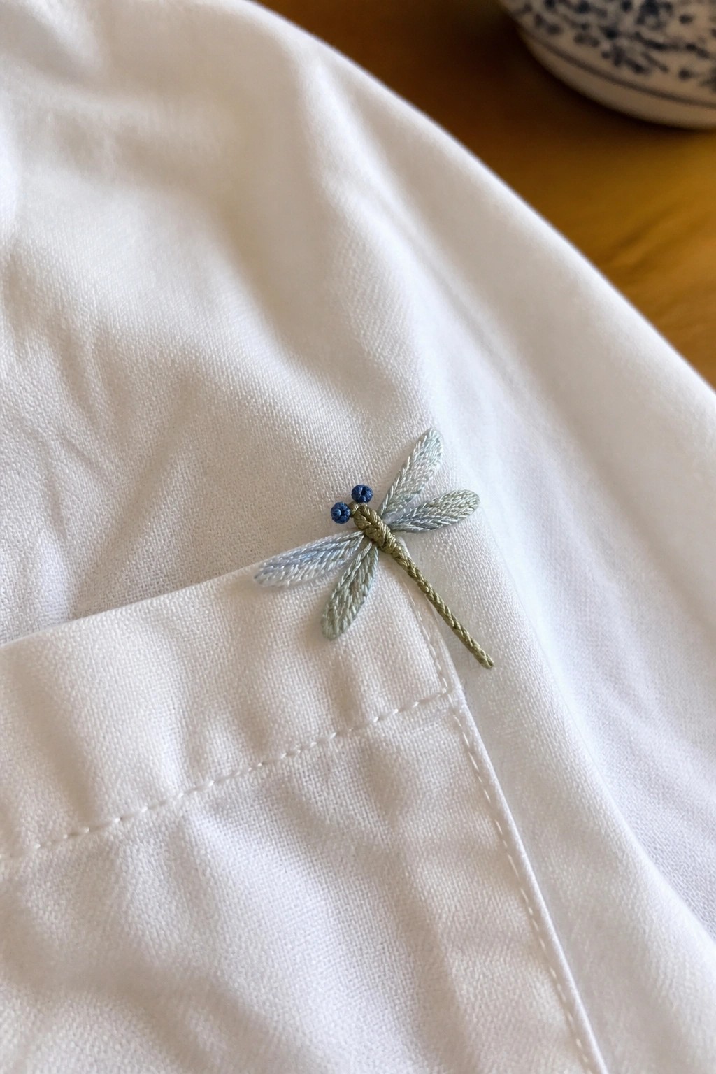 A dragonfly embroidered on the pocket of a white shirt using light blue and green threads.