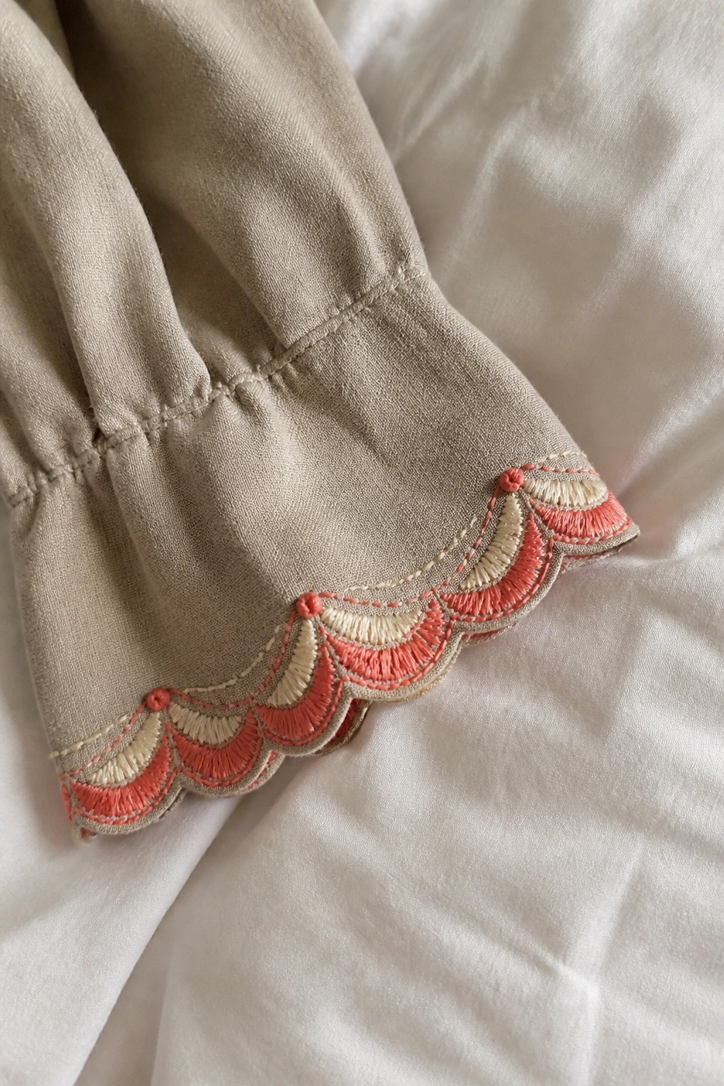 Beige sleeve cuff with coral and cream scalloped embroidery on white surface