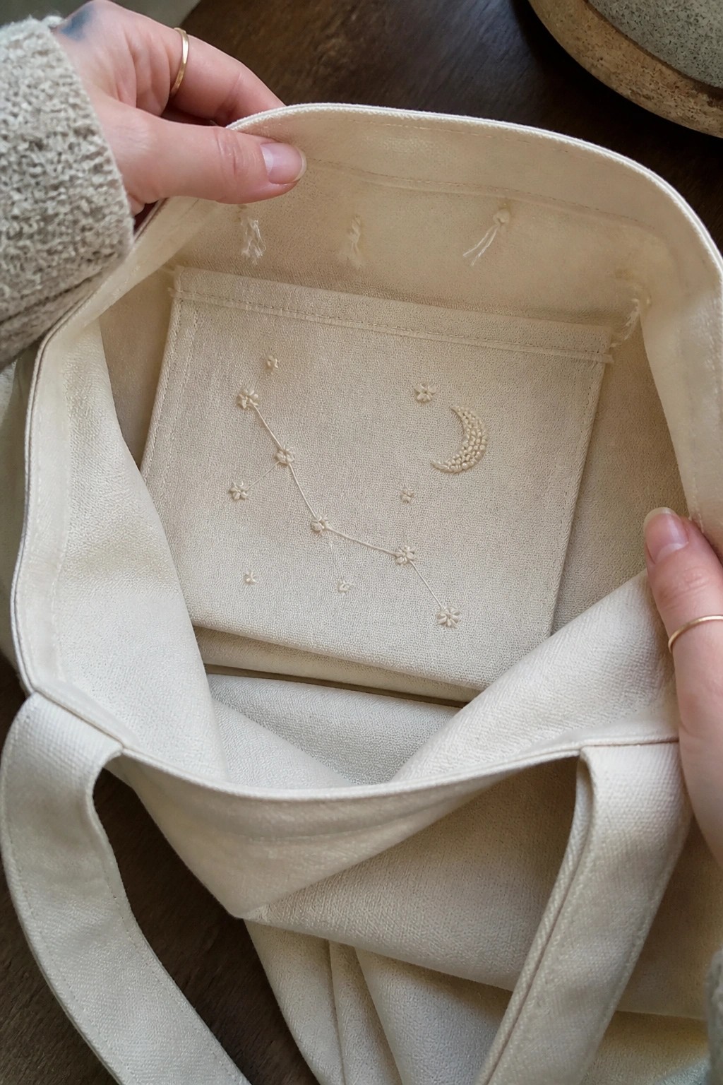 Hands opening cream tote bag with embroidered constellation and crescent moon inside pocket.