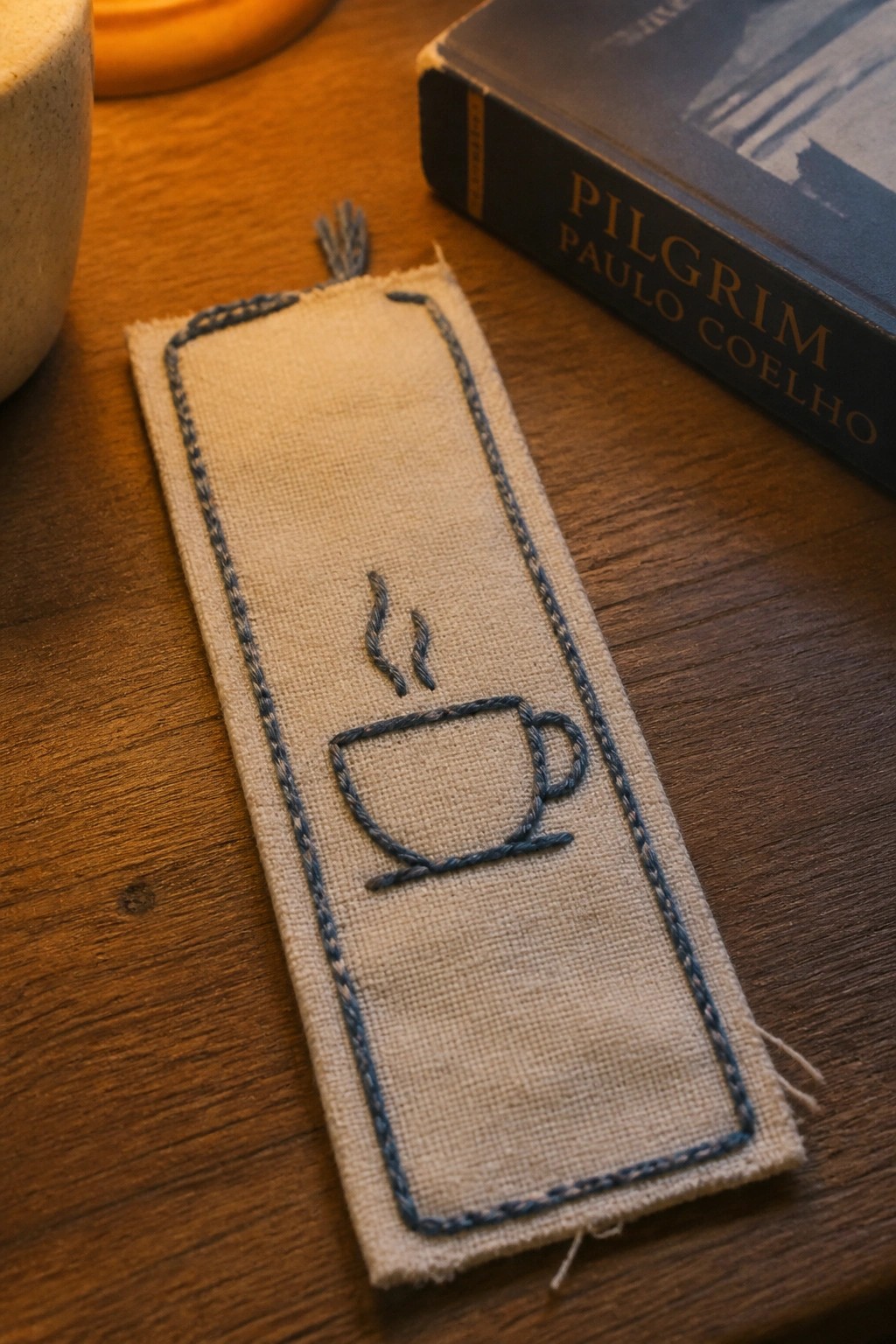 Embroidered beige bookmark with blue coffee cup design beside Paulo Coelho book on wood