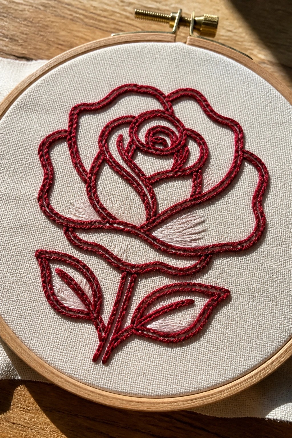 Red chain stitch rose with stem and leaves inside a wooden embroidery hoop on white fabric.