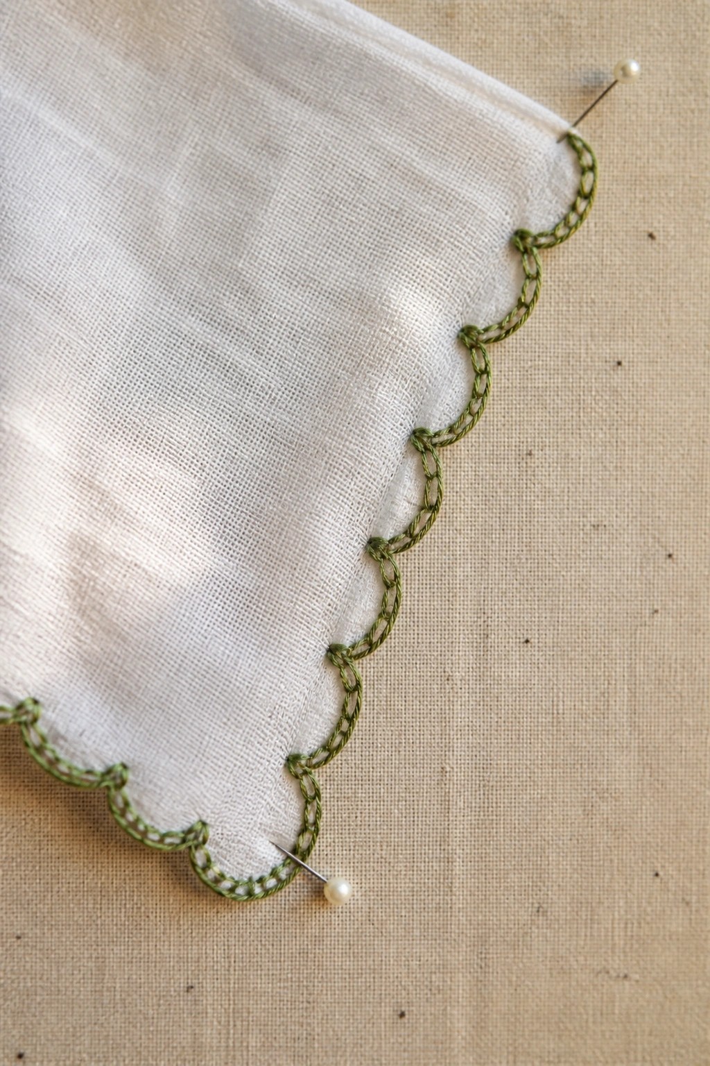 White fabric with green scalloped embroidery pinned by pearl-headed pins on beige cloth.