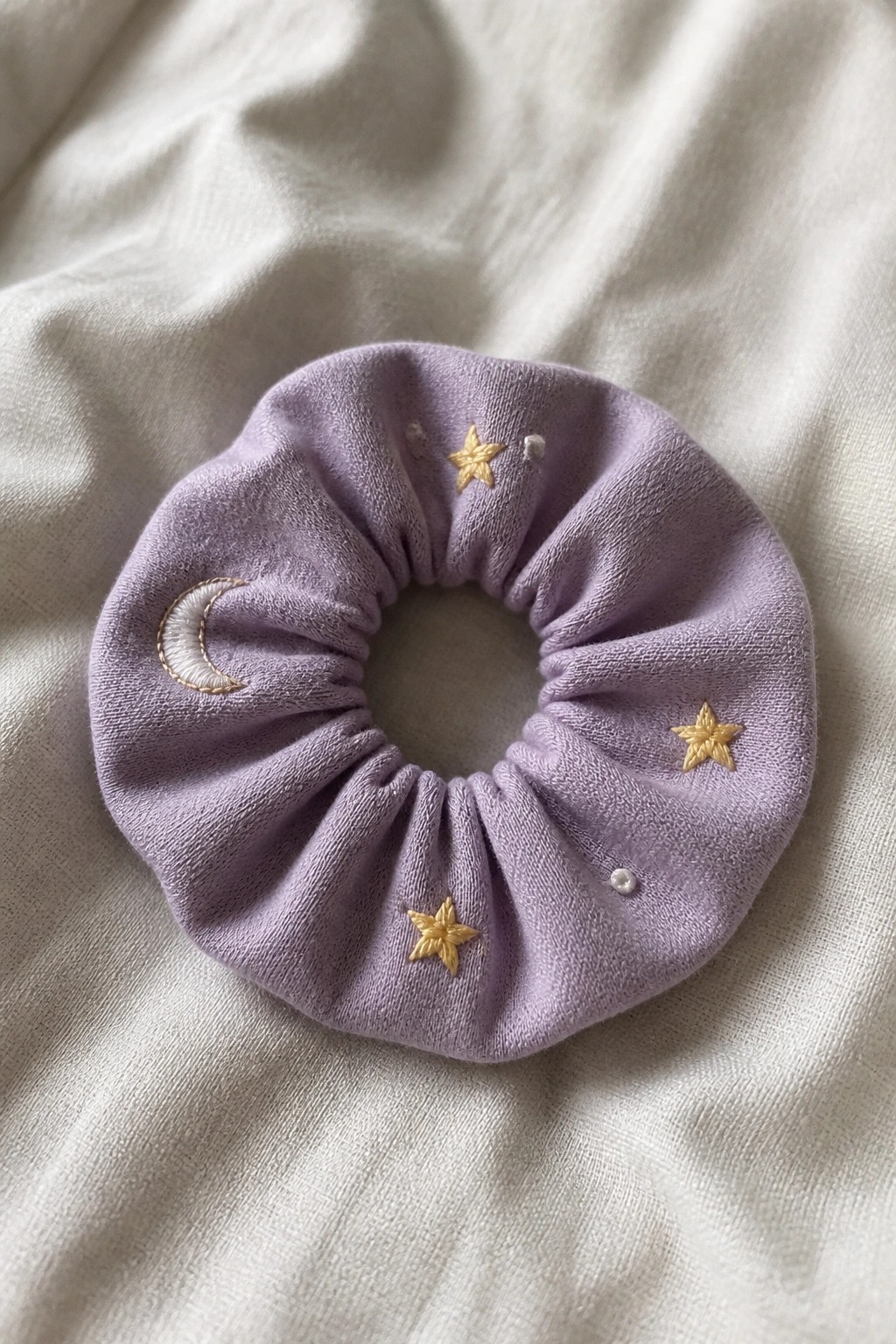 A lavender fabric scrunchie with a white crescent moon, several gold stars, and small white dots embroidered on the surface.