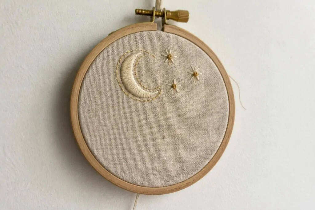 18 Cozy Embroidery Hoop Art Ideas Designed for Small Wall Displays Wooden hoop holding beige fabric embroidered with golden moon and white star flowers.