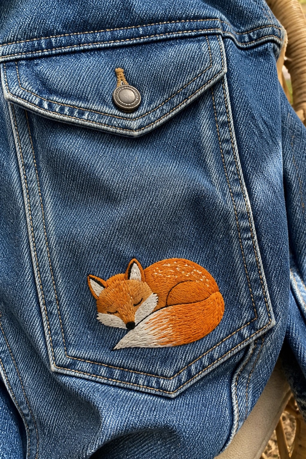 A sleeping orange fox is embroidered on the pocket of a blue denim jacket.